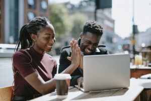 Two people sitting at an outdoor table using a laptop. They are smiling and doing a high-five, as if celebrating.