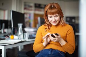 Happy young woman sitting relaxed at her desk and reading text message on her cell phone. Businessman using her mobile phone in office.