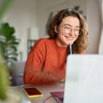 Happy young person using laptop sitting at desk writing notes while watching webinar, studying online, looking at pc screen learning web classes or having virtual call meeting remote working from home.