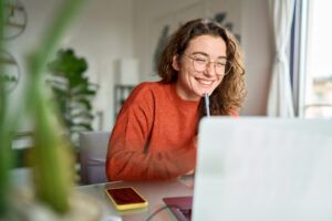 Happy young person using laptop sitting at desk writing notes while watching webinar, studying online, looking at pc screen learning web classes or having virtual call meeting remote working from home.