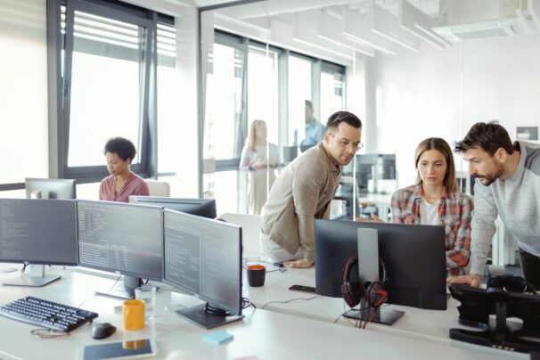 An office space with one person working on a computer monitor on the left and three people gathered around a computer monitor on the right. In the foreground, there is a three monitor set up with data software displayed.