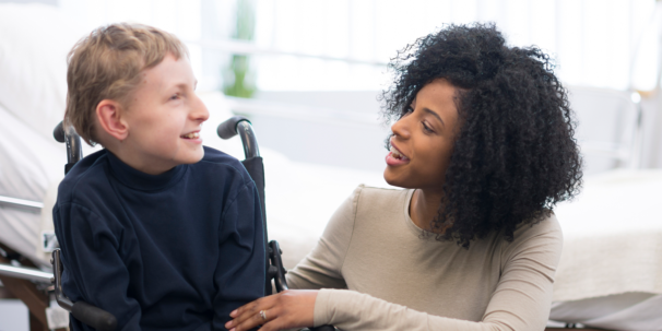 A woman smiling and talking to a young boy in a wheelchair.