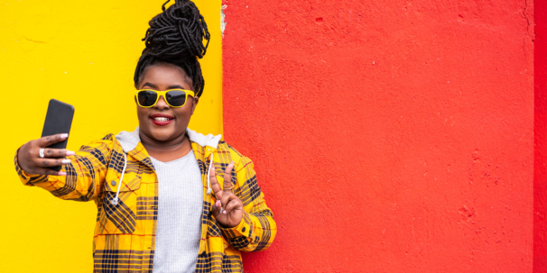 Woman in a bright yellow jacket and sunglasses taking a selfie against a colourful wall.