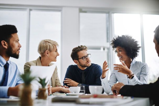 A group of professionals seated a table in an office looking at a colleague who is speaking.