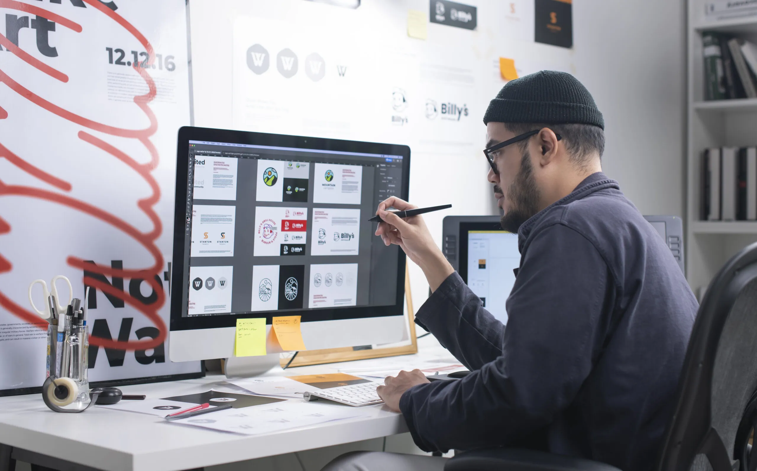 Individual sitting at a desk reviewing graphic designs on a computer monitor.