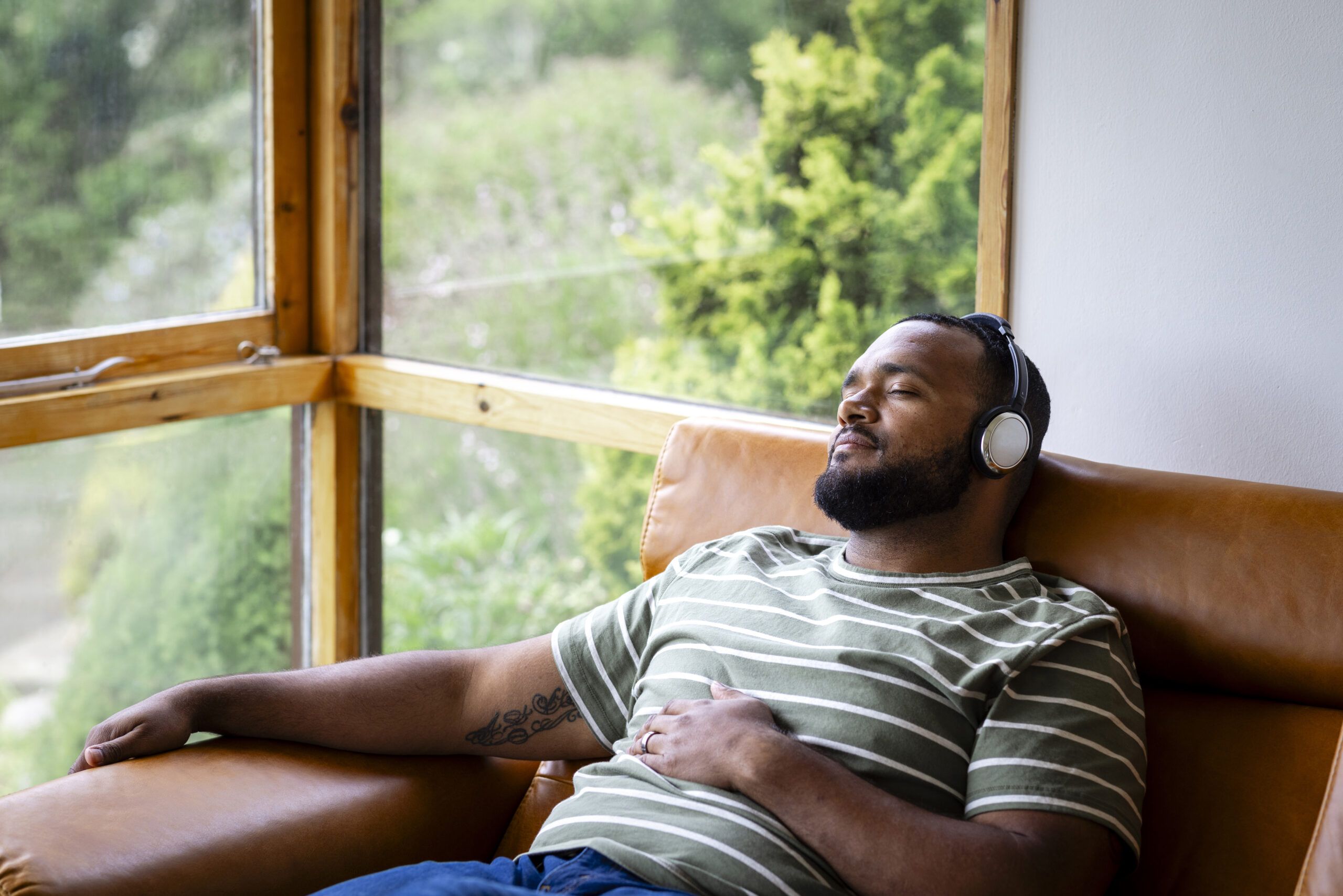 A mid adult man listening to music through wireless headphones and sitting back on a leather sofa in his living room at home in Hexham, North East England. He is listening to the music while looking content with his eyes closed.
