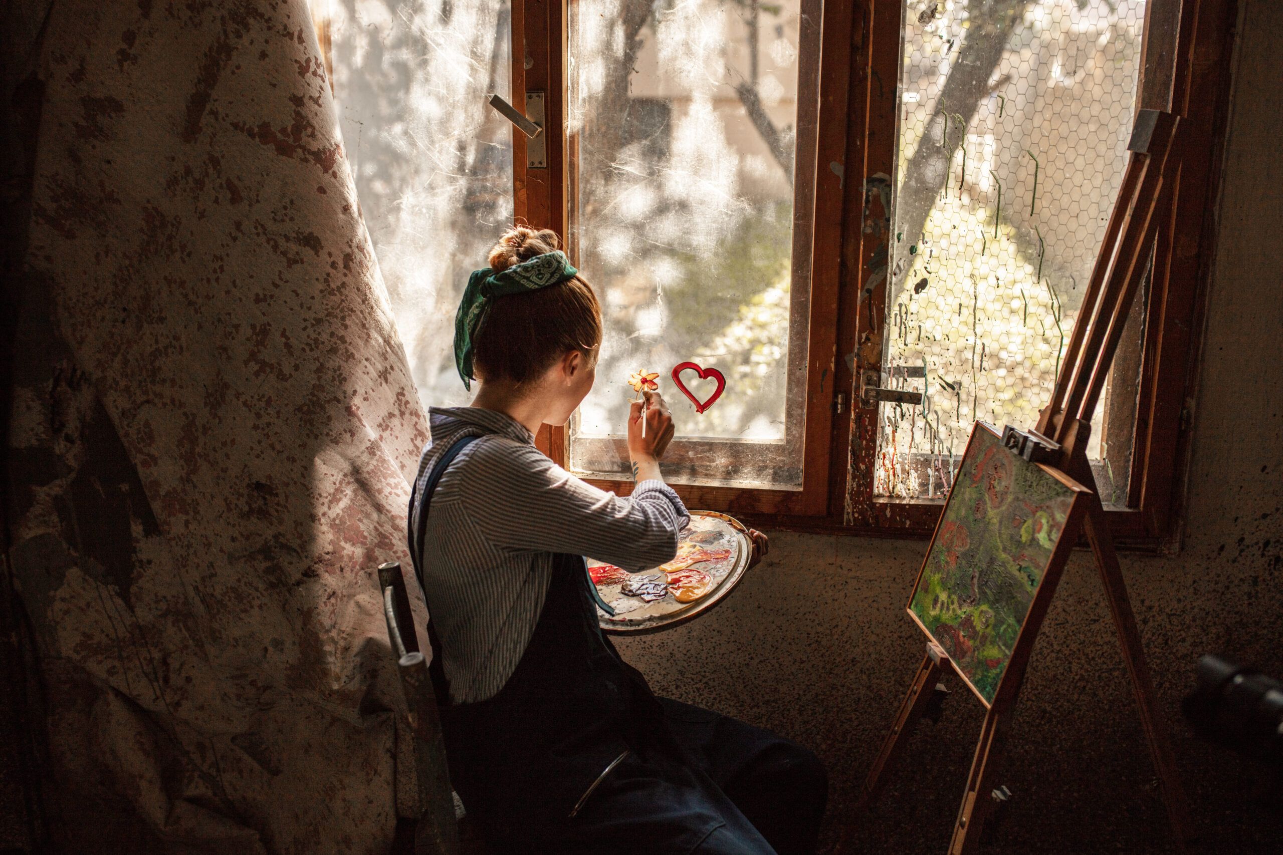 Individual sitting at an easel with a painting on, painting a flower on the window.