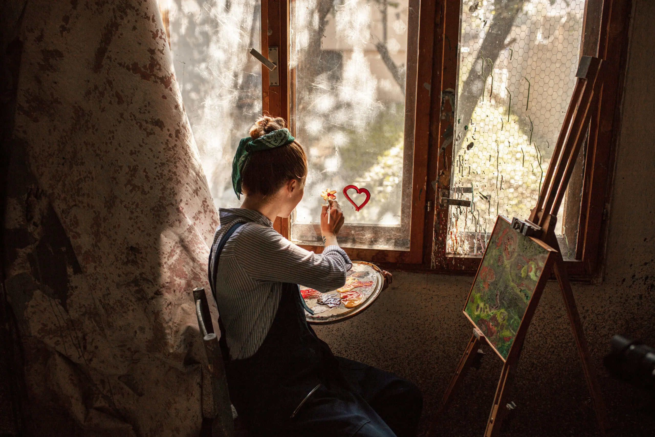 Individual sitting at an easel with a painting on, painting a flower on the window.