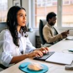 Two people work in a modern office setting. One is typing on a keyboard while the other is using a smartphone. They focus on their tasks in a bright indoor environment.
