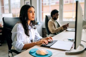 Two people work in a modern office setting. One is typing on a keyboard while the other is using a smartphone. They focus on their tasks in a bright indoor environment.