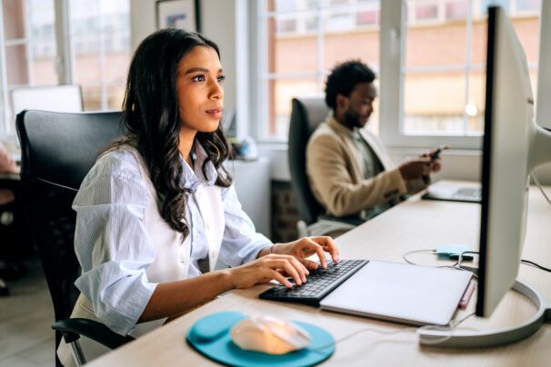 Two people work in a modern office setting. One is typing on a keyboard while the other is using a smartphone. They focus on their tasks in a bright indoor environment.
