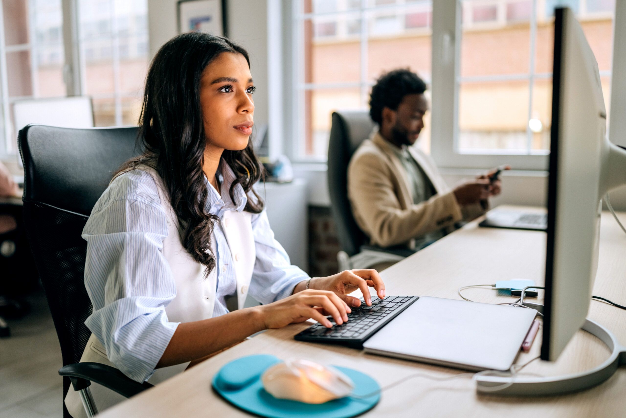 Two people work in a modern office setting. One is typing on a keyboard while the other is using a smartphone. They focus on their tasks in a bright indoor environment.