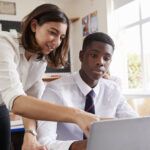Female Teacher Helping Pupil Using Computer In Classroom